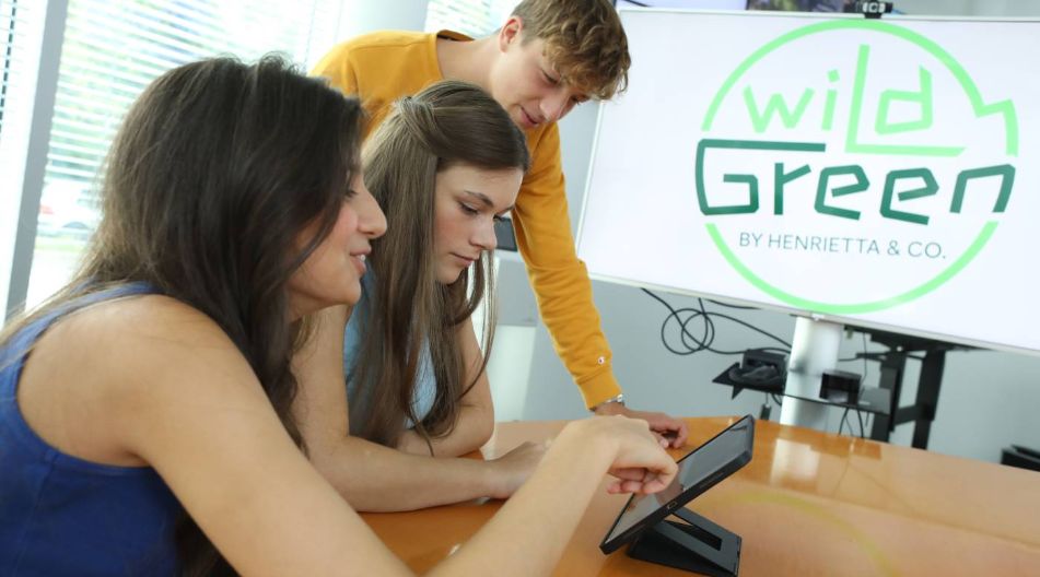 Drei junge Menschen (zwei Frauen und ein Mann) schauen auf ein Tablet, das auf einem Tisch steht. Im Hintergrund ist ein Bildschirm mit dem Logo "wild Green by Henrietta & Co." sichtbar.