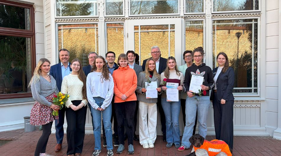 Gruppe von Jugendlichen und Erwachsenen vor einem Gebäude, einige halten Urkunden und Blumen; ein Hund mit Warnweste liegt vorne.