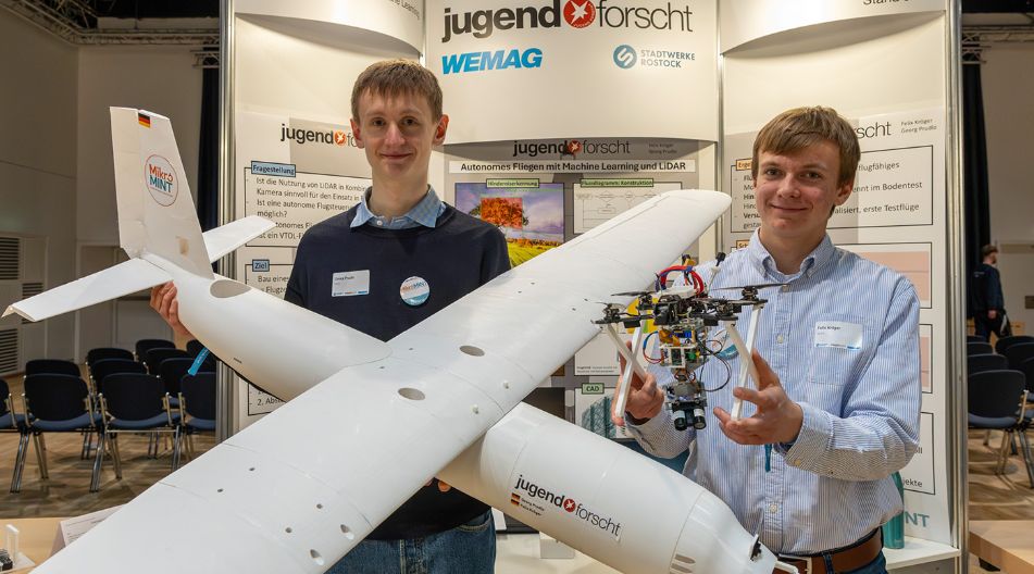 Zwei junge Männer präsentieren ein selbstgebautes Flugzeug und eine Drohne auf einer Technikmesse.